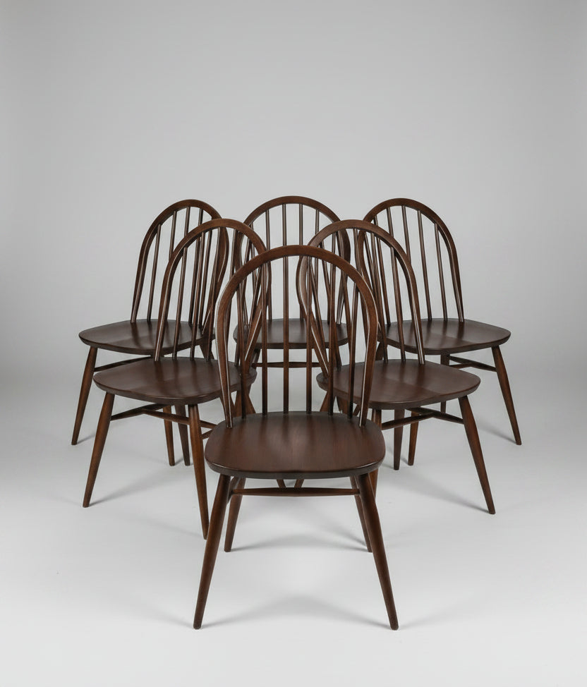 Six dark brown wooden spindle-back dining chairs arranged in a group on a worn reddish-brown floor inside what appears to be a furniture store or warehouse.
