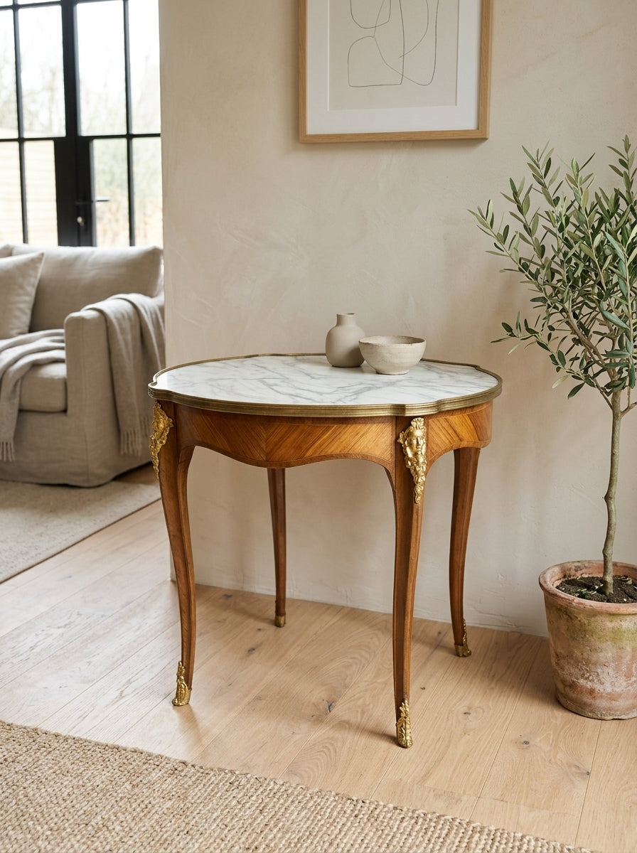A round console table with a white marble top and a warm brown wood frame with gold-tone legs, styled in a bright, modern living room next to a potted olive tree.