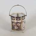 Two decorative metal and glass canisters with ornate silver-tone handles and lids, one featuring a white and blue floral pattern and the other a blue panel with white classical figures, set against a plain light background.