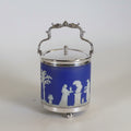 Two decorative metal and glass canisters with ornate silver-tone handles and lids, one featuring a white and blue floral pattern and the other a blue panel with white classical figures, set against a plain light background.