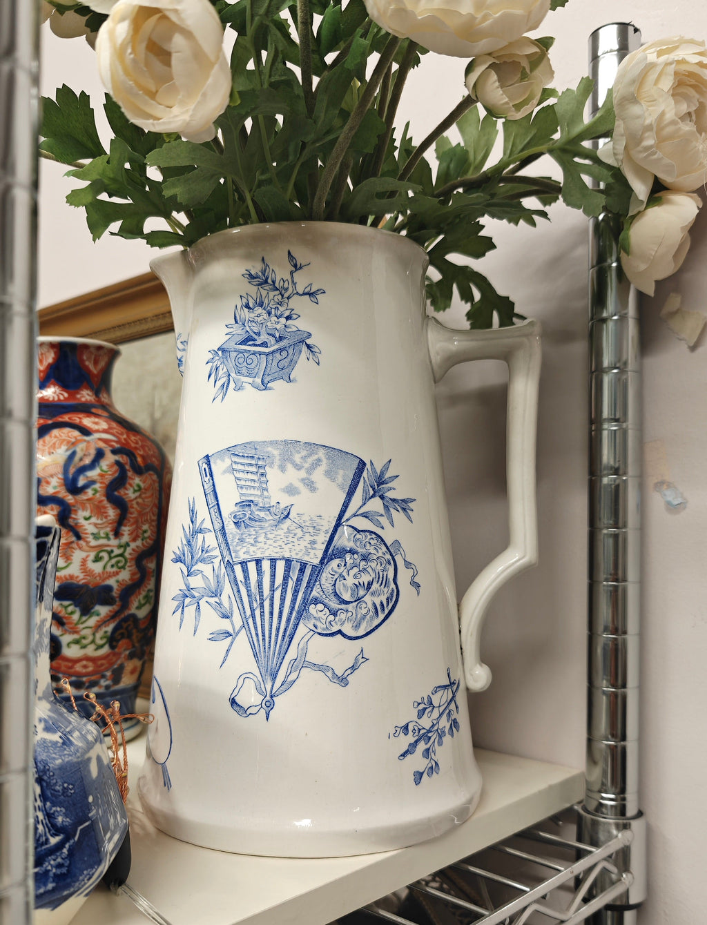 A white ceramic pitcher with blue toile-style illustrations, filled with cream-colored peony flowers, displayed on a white shelf in a wire shelving unit.