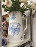A white ceramic pitcher with blue toile-style illustrations, filled with cream-colored peony flowers, displayed on a white shelf in a wire shelving unit.