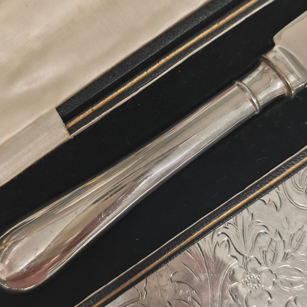 A close-up of a polished silver-toned metal knife handle resting inside an open black presentation box lined with cream-colored fabric, placed on a textured silver surface.