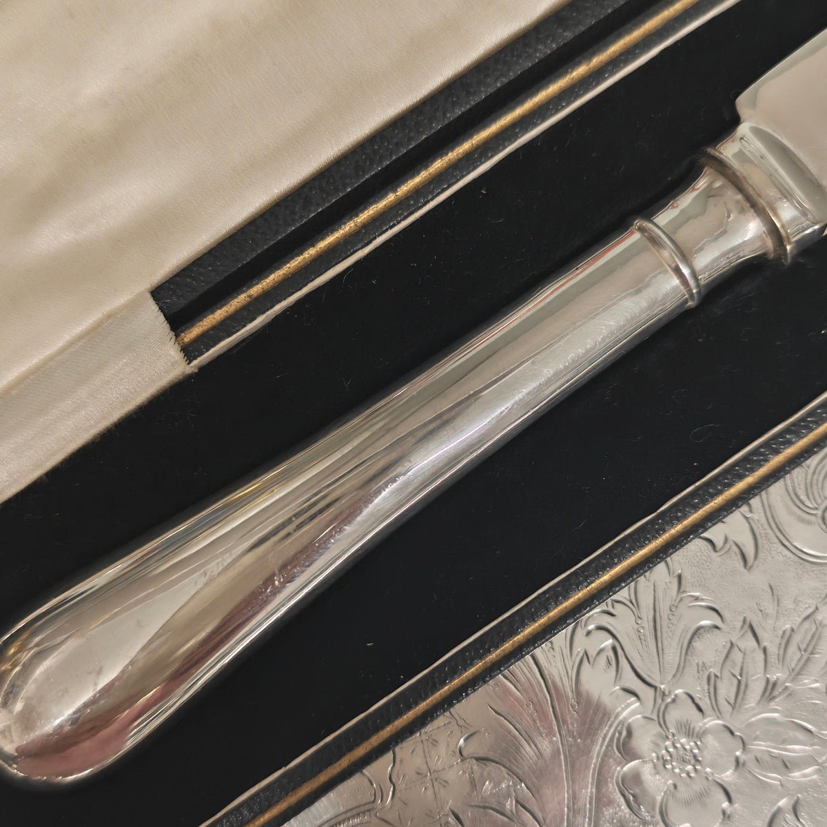 A close-up of a polished silver-toned metal knife handle resting inside an open black presentation box lined with cream-colored fabric, placed on a textured silver surface.