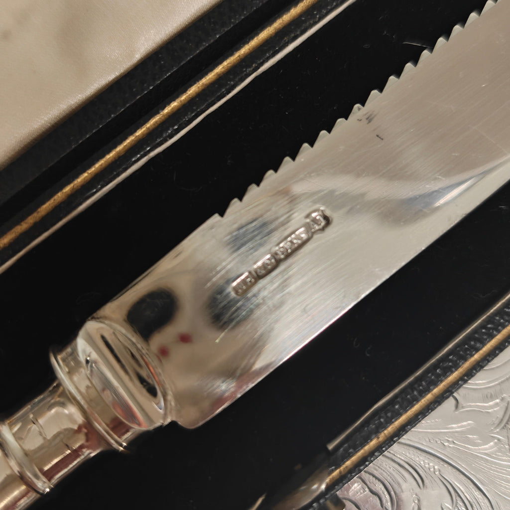 A close-up of a silver serrated knife with a decorative handle, resting inside a black presentation box with gold trim.