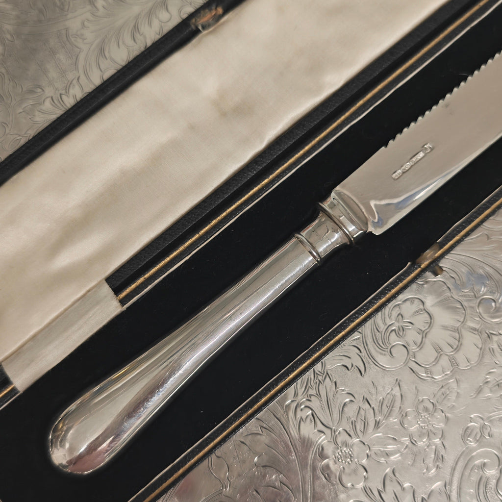 A silver-toned metal knife with a polished handle, resting inside an open black presentation box lined with cream fabric, placed on a textured silver surface.