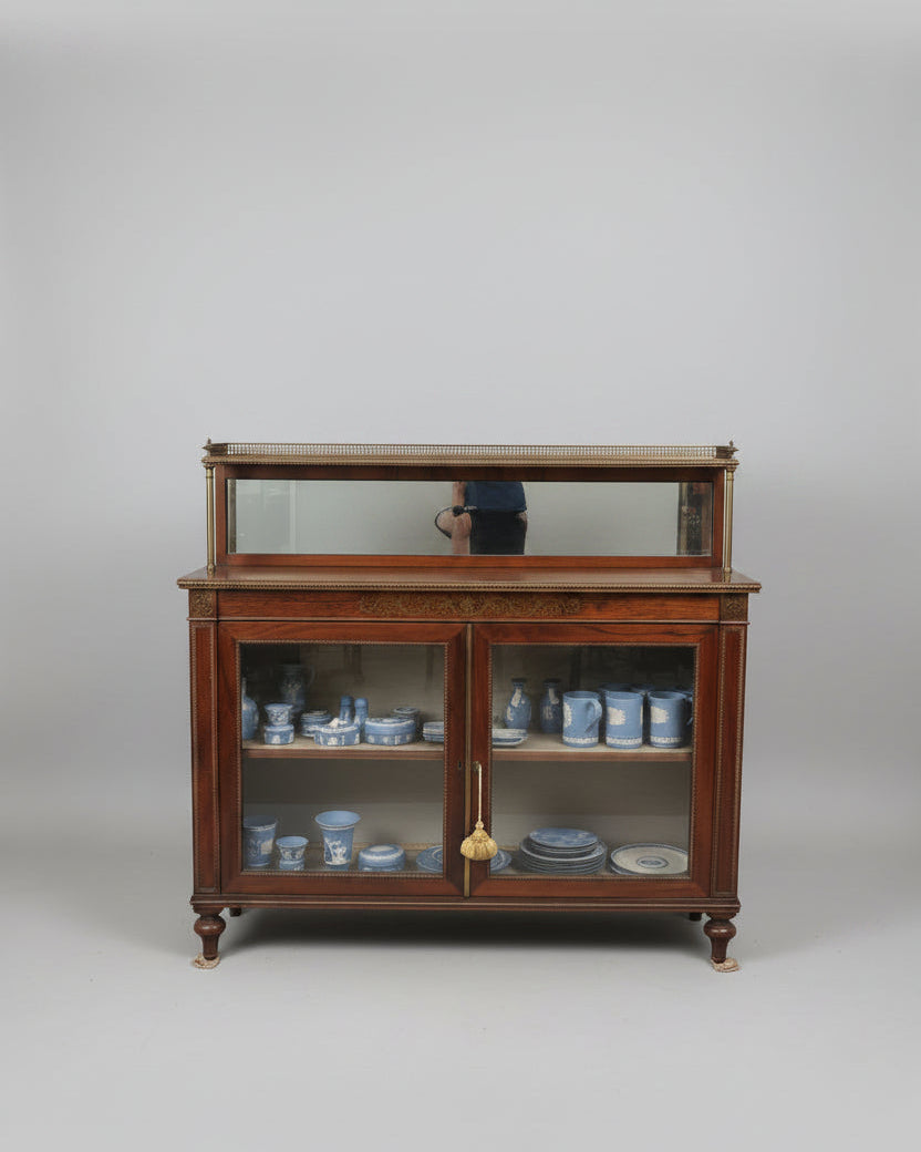 A dark wood china cabinet with glass doors in an antique shop, with a woman pointing at the cabinet top display.