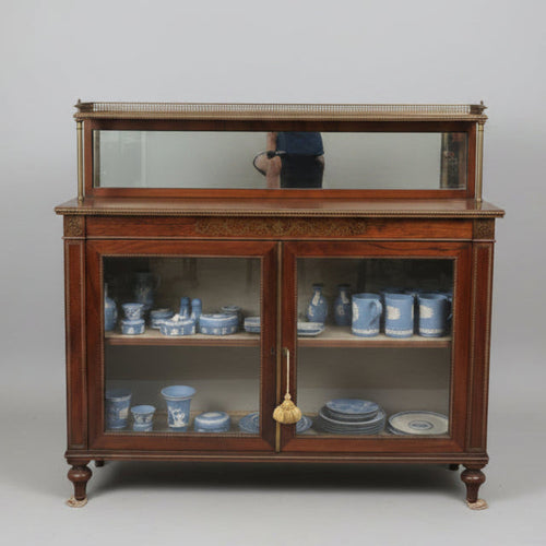 A dark wood china cabinet with glass doors in an antique shop, with a woman pointing at the cabinet top display.