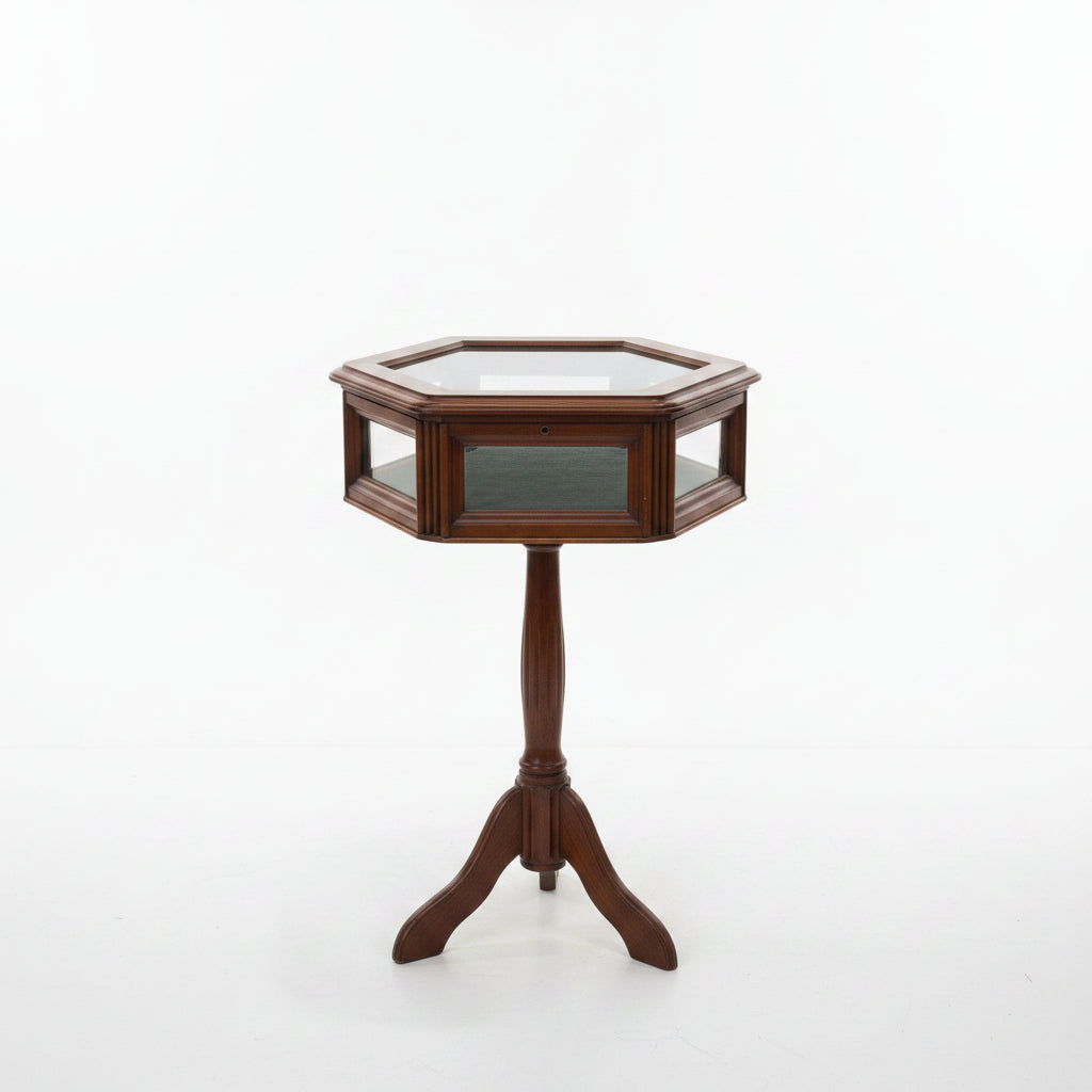 A hexagonal wooden display table with a glass top and three legs, photographed against a plain wall and light wood floor.