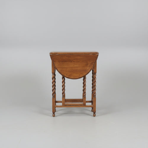 A dark brown wooden dressing table with a drop-leaf top, twisted legs, a shelf, and an attached storage chest, photographed in a furniture showroom.