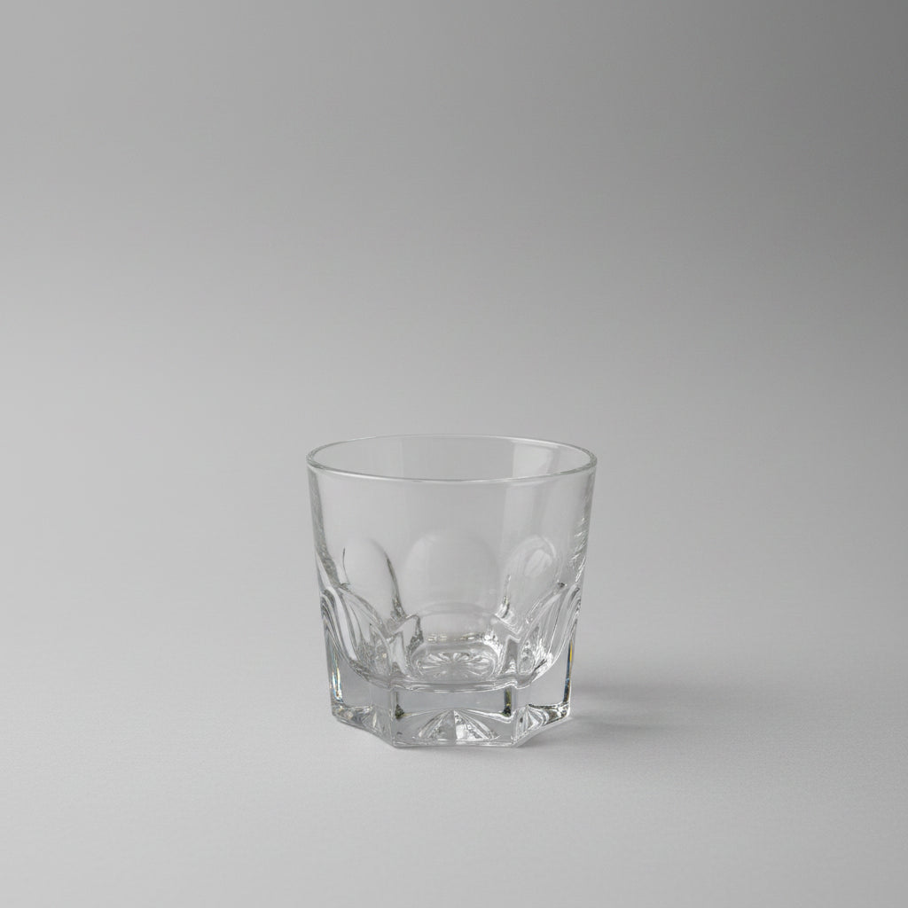A clear cut-glass rocks tumbler with a faceted base, shown on a dark wooden table.