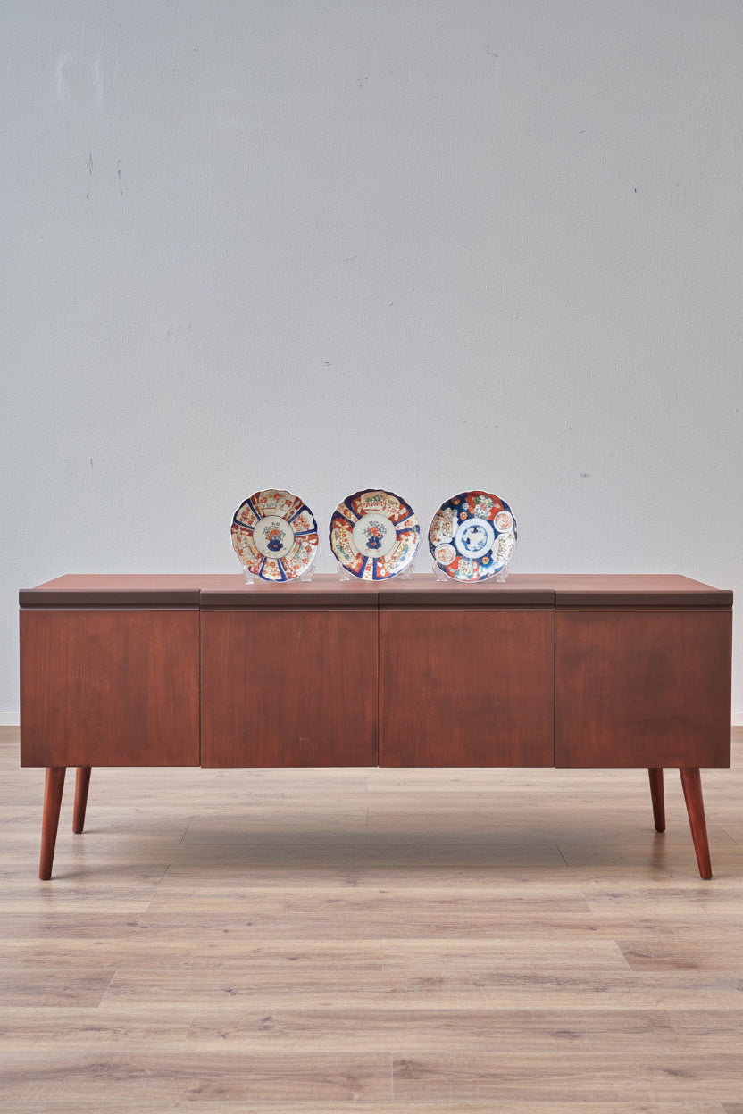 A mid-century modern wooden sideboard console table with a warm brown finish, styled on a red patterned rug with decorative plates on top, set against a light gray wall.