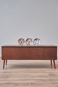 A mid-century modern wooden sideboard console table with a warm brown finish, styled on a red patterned rug with decorative plates on top, set against a light gray wall.