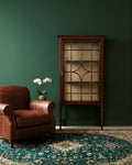 Brown armchair and wooden display cabinet against a green wall with a patterned rug.
