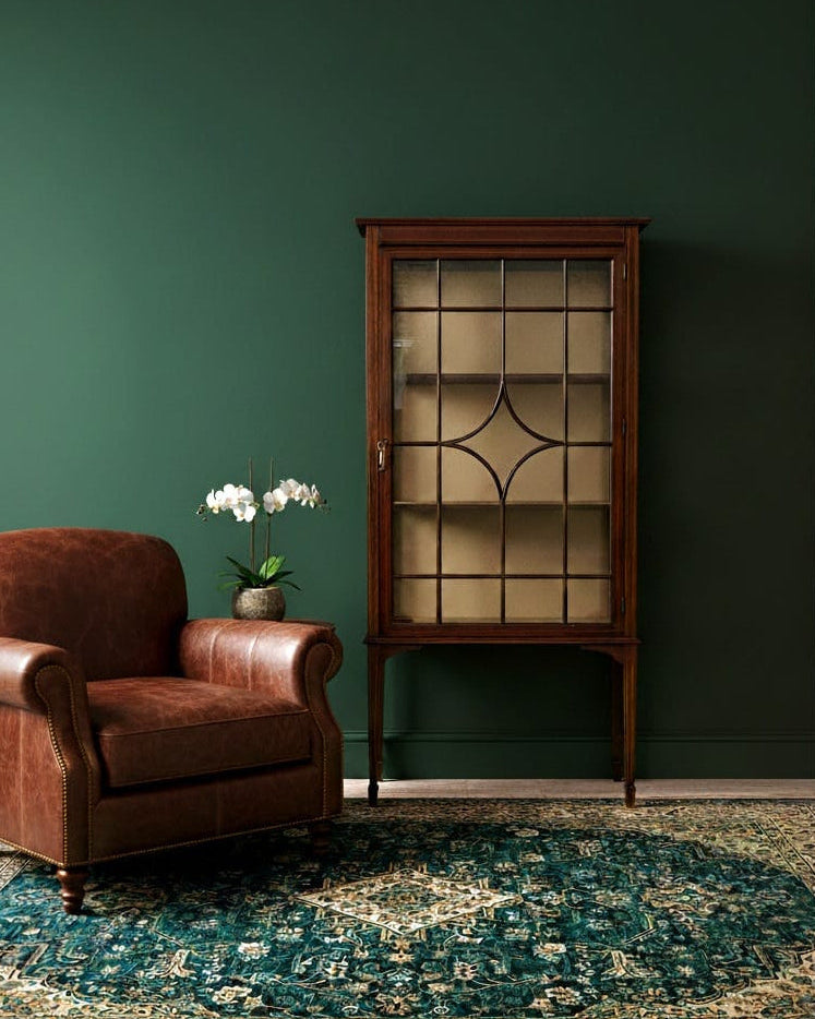 Brown armchair and wooden display cabinet against a green wall with a patterned rug.