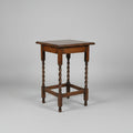 A small square wooden table with turned legs and a lower support rail, photographed on a worn reddish-brown floor in what appears to be a shop or storage area.