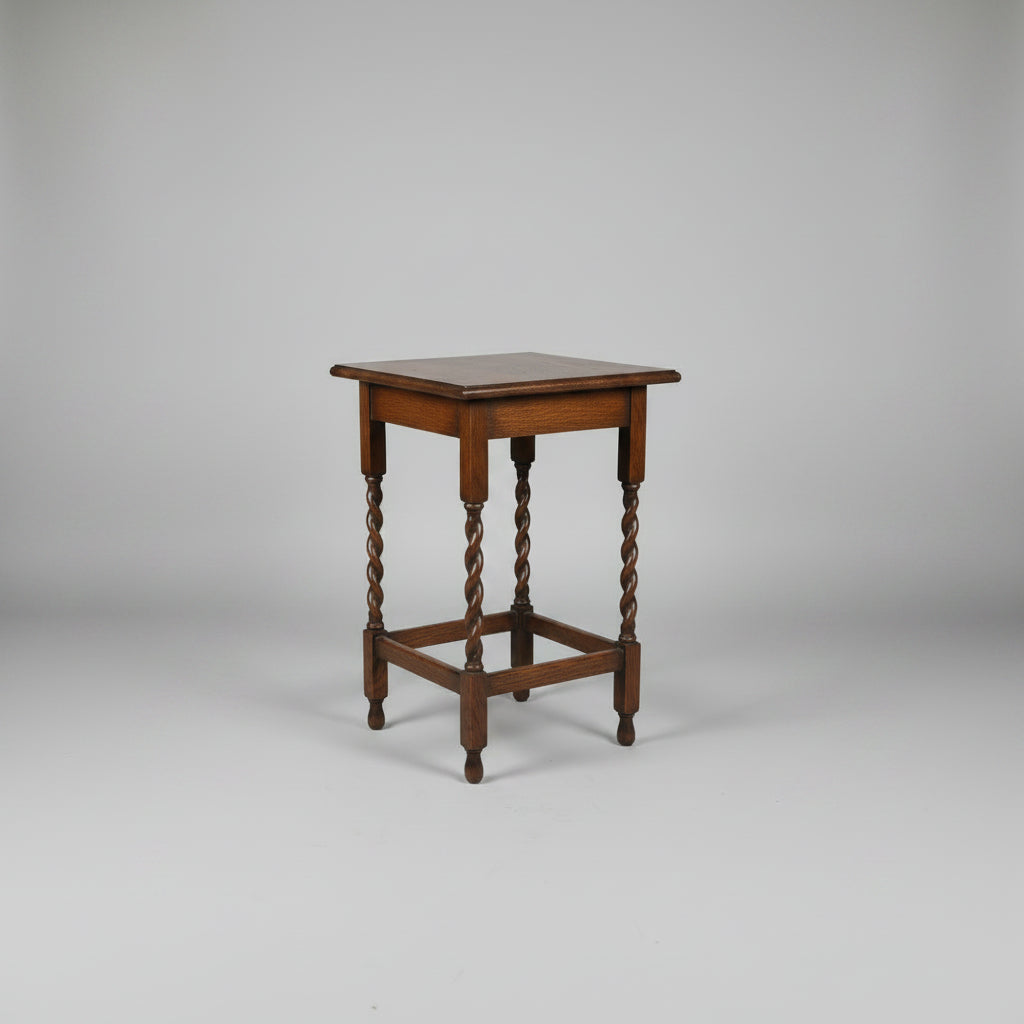 A small square wooden table with turned legs and a lower support rail, photographed on a worn reddish-brown floor in what appears to be a shop or storage area.