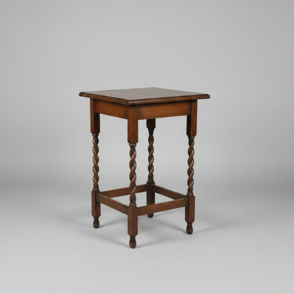A small square wooden table with turned legs and a lower support rail, photographed on a worn reddish-brown floor in what appears to be a shop or storage area.