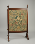 An antique wooden tapestry stand featuring a detailed floral and pastoral tapestry panel in the center, set against a neutral gray studio background.