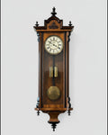 A traditional wall-mounted grandfather clock with a wooden case and a white clock face, standing above a shelf with a plant and some decorative items.