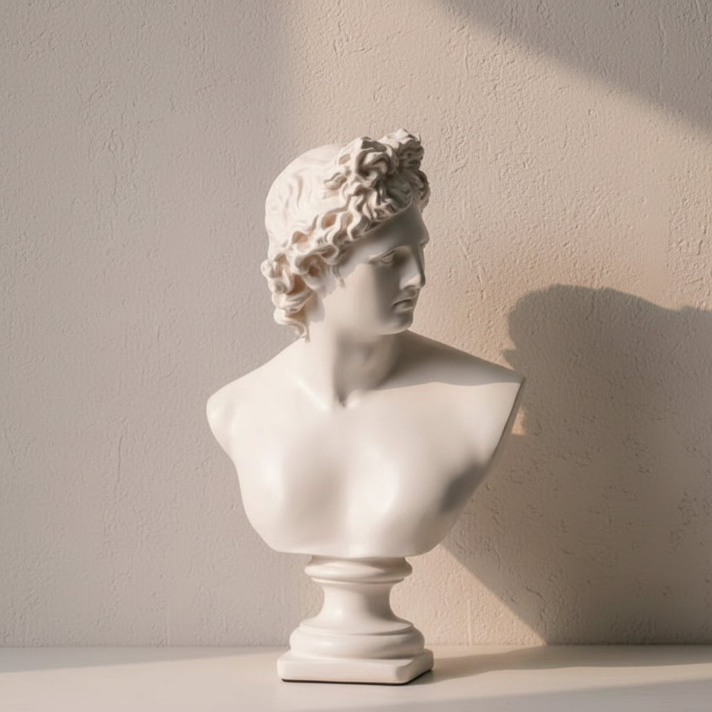 A white classical-style bust sculpture displayed indoors against a dark textured wall, with draped beige fabric, a clipboard, and books in the background.