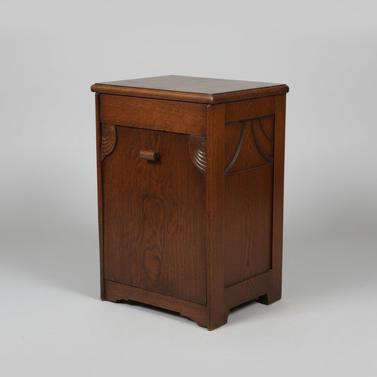 A vintage dark brown wooden chest or cabinet with a single front drawer and a small round knob, photographed in a room with a worn multicolored floor and other furniture partially visible.
