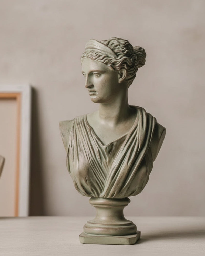 A classical-style stone bust sculpture of a draped female figure, displayed against a dark textured wall with vintage sheet music and draped fabric in the background.