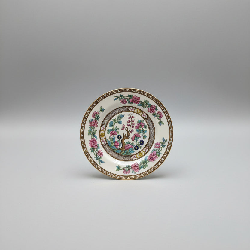 A round white porcelain plate with a colorful floral and bird design, displayed on a dark brown leather surface with gold detailing.