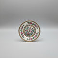 A round white porcelain plate with a colorful floral and bird design, displayed on a dark brown leather surface with gold detailing.