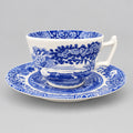 A classic blue and white porcelain teacup and saucer set with a detailed landscape print, displayed on a wooden table against a light wall.
