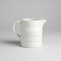 A white ceramic milk jug with a handle, placed on a lace tablecloth.
