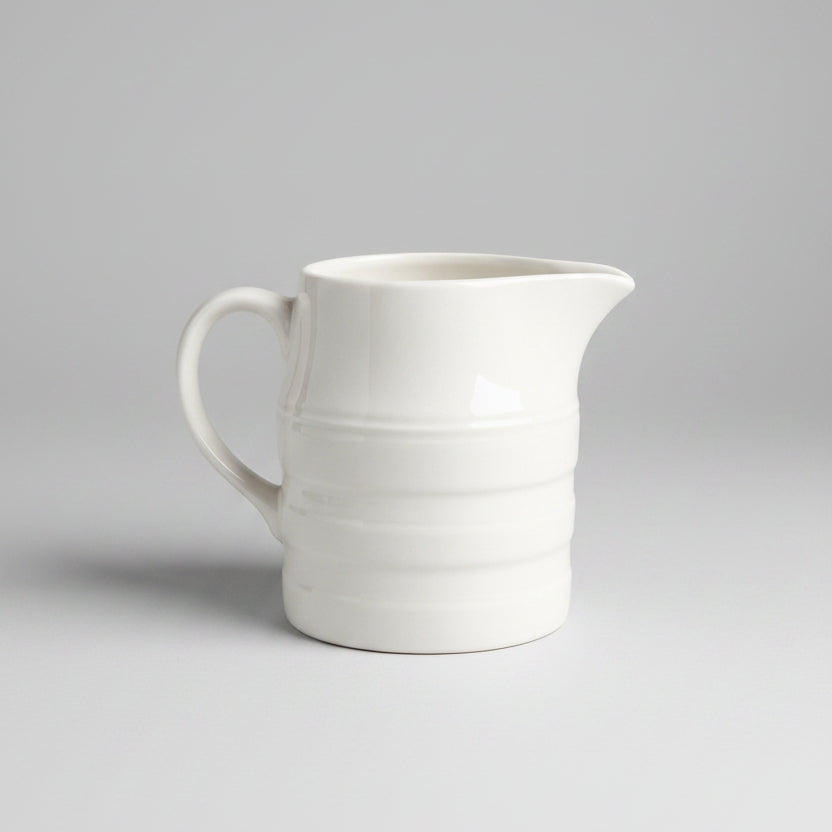 A white ceramic milk jug with a handle, placed on a lace tablecloth.