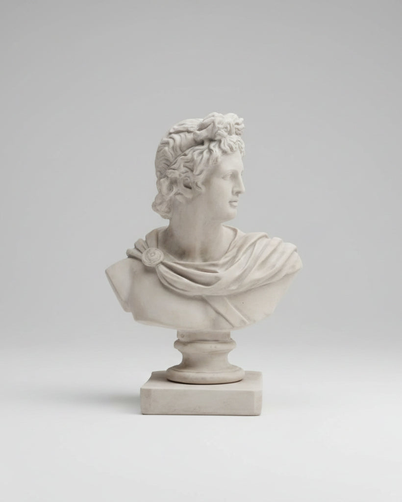 A classical-style stone bust sculpture of a draped male figure in profile, displayed on a pedestal base against an indoor paneled wall background.