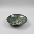 A large round ceramic bowl with a glossy, swirling wood-grain pattern in shades of green, gray, and black, displayed on a wooden table in what appears to be a furniture showroom.
