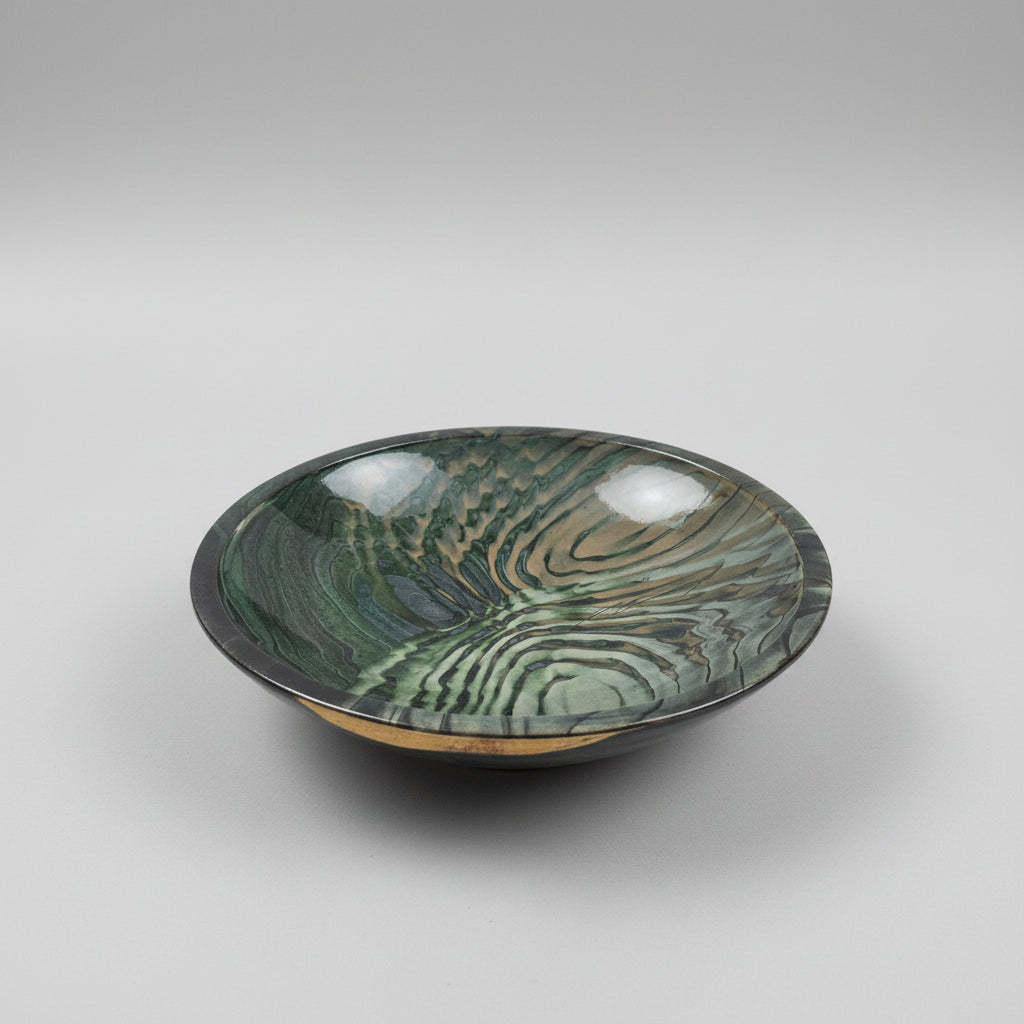 A large round ceramic bowl with a glossy, swirling wood-grain pattern in shades of green, gray, and black, displayed on a wooden table in what appears to be a furniture showroom.