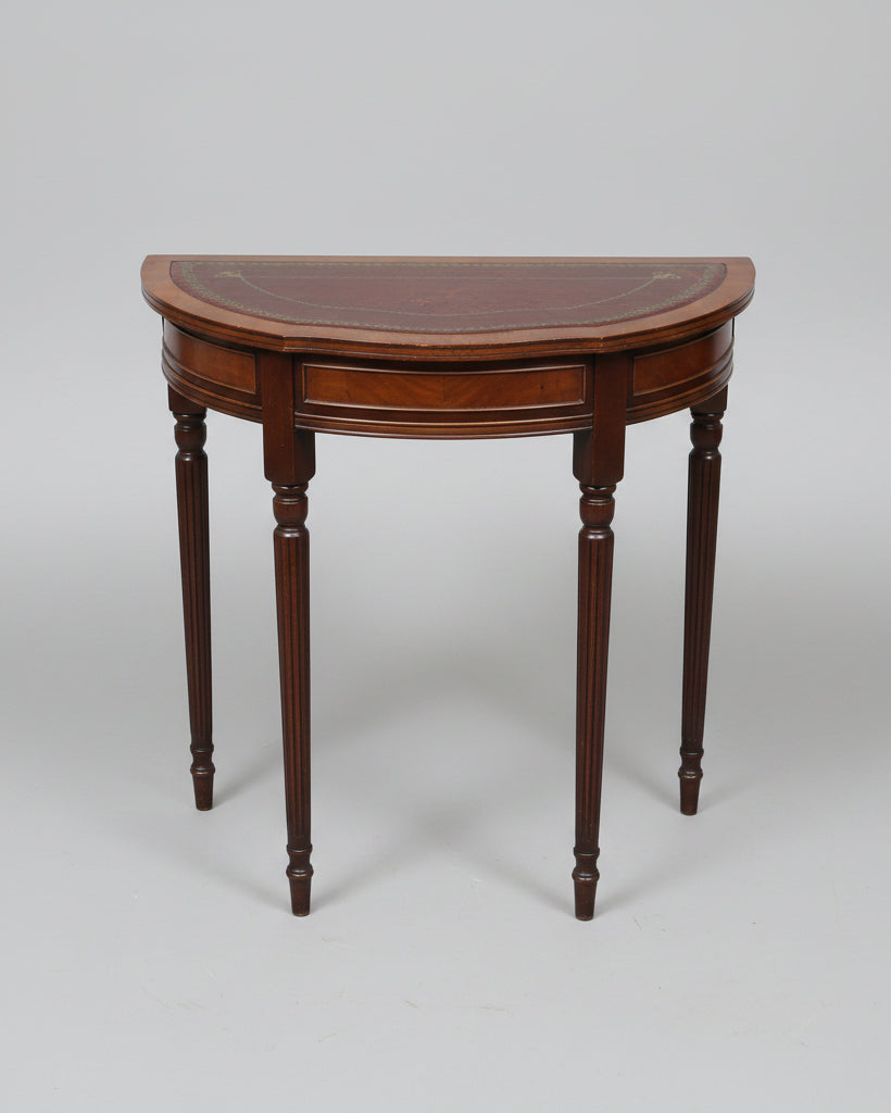 A vintage wooden half-round console table with a dark brown leather inlay and gold decorative border, shown in a cluttered storage or warehouse setting.