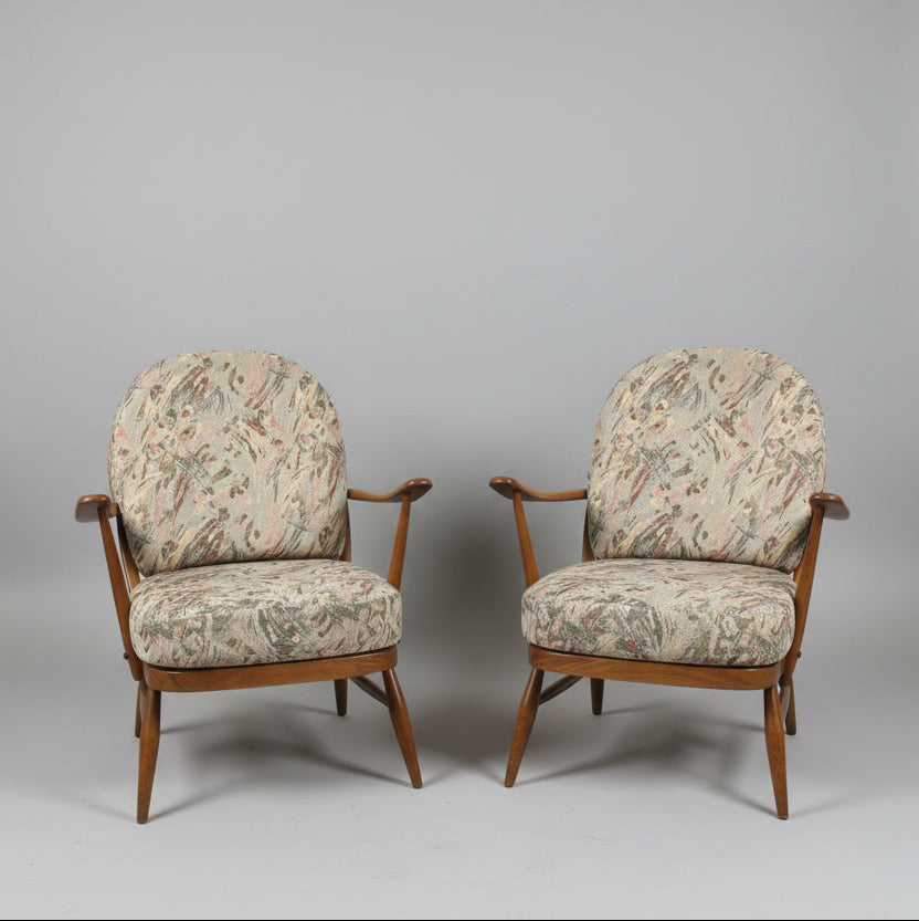 Two vintage wooden armchairs with patterned beige and brown upholstery, shown in a warehouse or showroom setting with a red door and red floor.