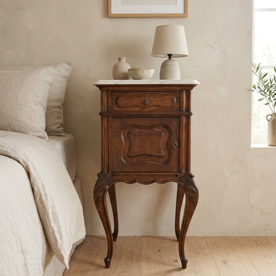 A dark brown wooden nightstand with carved details and cabriole-style legs, styled in a bedroom next to a bed with beige linens. It features a white marble top, a single drawer with a round knob, and a central cabinet panel with a small lock.