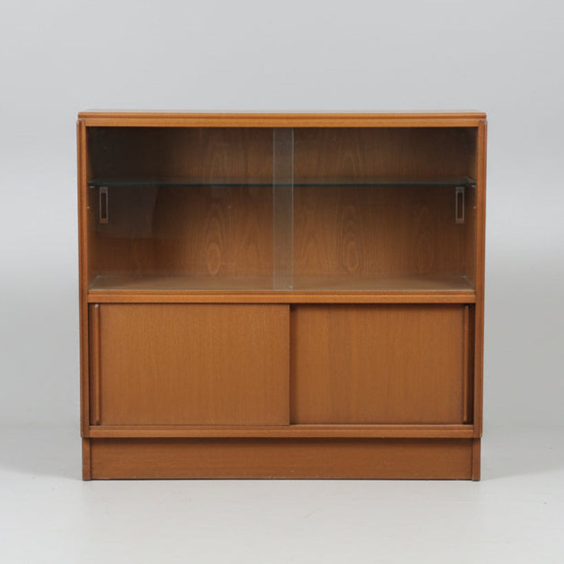 A mid-century modern wooden display cabinet with glass doors and two glass shelves, photographed in what appears to be a furniture showroom or warehouse.