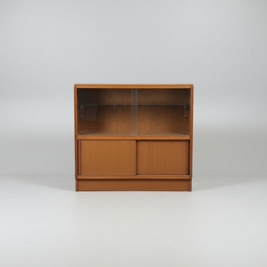 A mid-century modern wooden display cabinet with glass doors and two glass shelves, photographed in what appears to be a furniture showroom or warehouse.