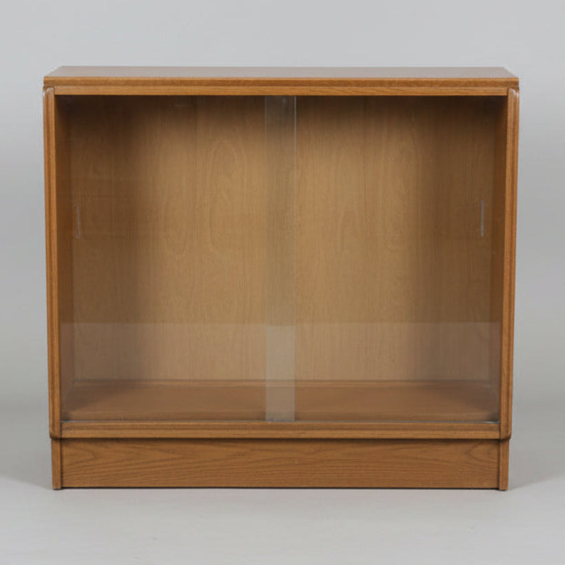 A mid-century wooden display cabinet with two glass sliding doors, shown in what appears to be a furniture showroom or warehouse.