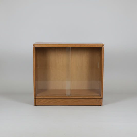 A mid-century wooden display cabinet with two glass sliding doors, shown in what appears to be a furniture showroom or warehouse.