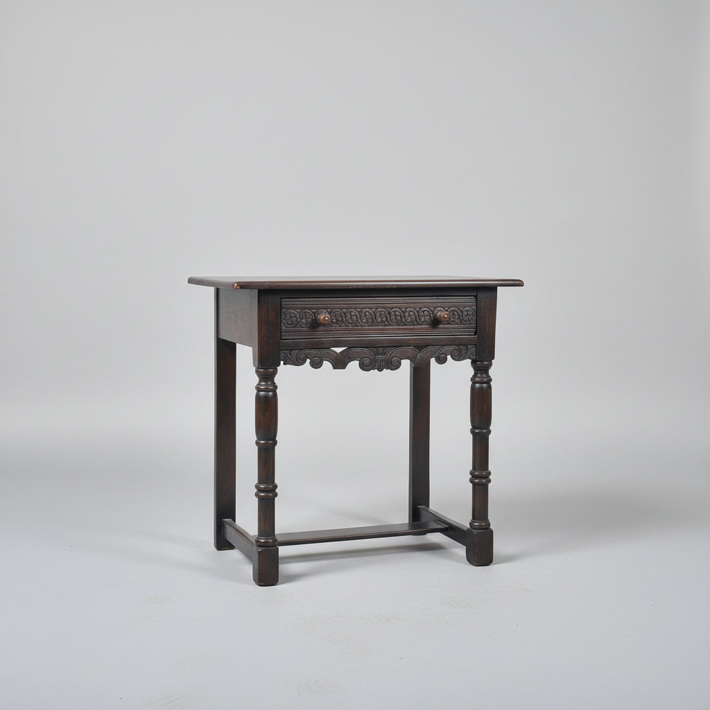 Wooden side table with a drawer on a red patterned rug against a gray wall.