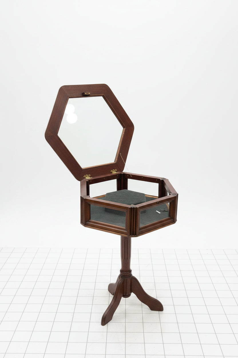 A hexagonal wooden display case or cabinet with an open hinged lid, glass panels, brass hinges, and a dark green velvet interior, shown on a light gray wood floor against a white wall.