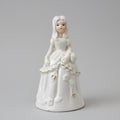 A white porcelain figurine of a young girl with long light hair, wearing a long dress and holding a bouquet, displayed on a dark wooden surface against a textured off-white wall.