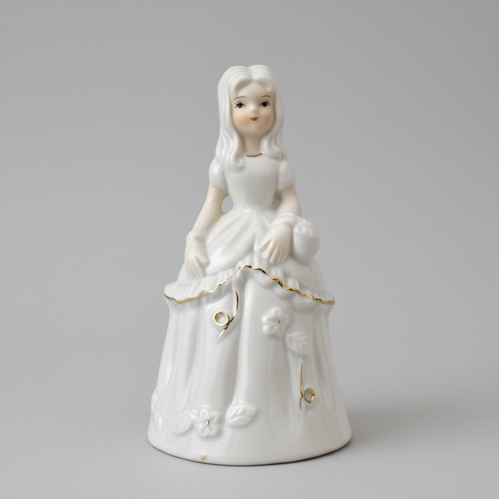 A white porcelain figurine of a young girl with long light hair, wearing a long dress and holding a bouquet, displayed on a dark wooden surface against a textured off-white wall.