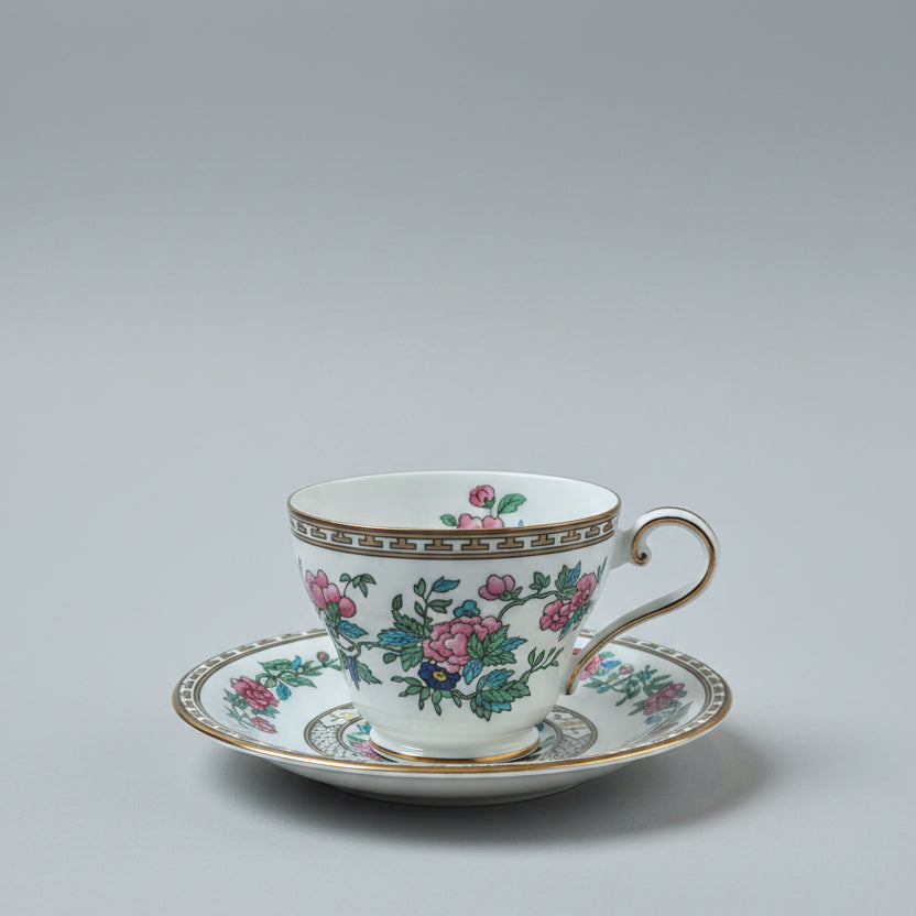 A white porcelain teacup and saucer set with a pink floral pattern and gold trim, resting on a wooden table with a wooden chair in the background.