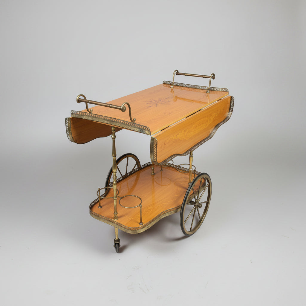 A vintage wooden serving cart with a polished light oak top, decorative metal trim, and brass-toned legs, photographed in a warehouse or showroom with red flooring and upholstered furniture in the background.