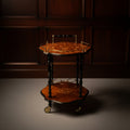An ornate two-tier wooden serving cart with a scalloped marquise-shaped top, featuring a floral inlay pattern, turned dark wood legs, and brass handles on the upper tier, set on a dusty red floor.