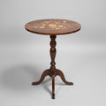 A round wooden pedestal table with an inlaid floral pattern on the tabletop, surrounded by other furniture in what appears to be a shop or storage area.